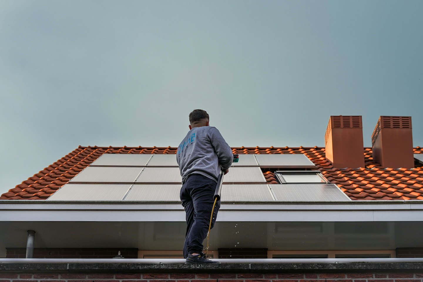 Mitarbeiter reinigt Solarmodule auf einem orange gefliesten Dach mit einer langen Bürste.